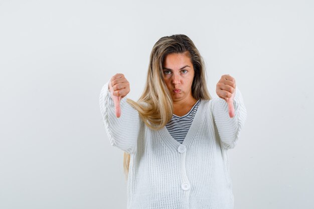 Blonde girl showing thumbs down with both hands in striped t-shirt, white cardigan and jean pants and looking displeased , front view.