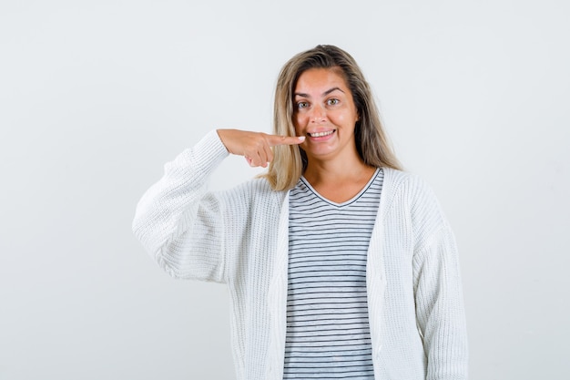 Free Photo blonde girl in striped t-shirt, white cardigan and jean pants pointing to teeth and looking happy , front view.