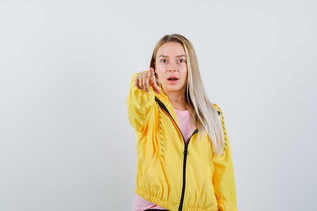 Blonde girl in t-shirt, jacket pointing at camera and looking amazed