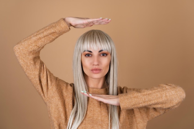 A blonde girl with bangs and brown daytime make-up in a sweater on a beige background pulls her hands above her head and under her chin