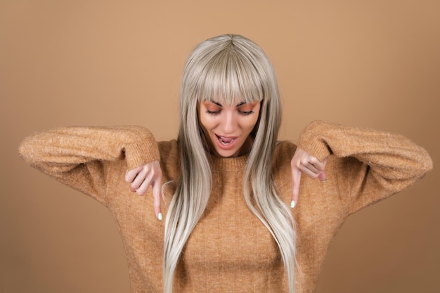 Free photo blonde girl with bangs and brown daytime makeup in a sweater on a beige background smiles excitedly points her finger down