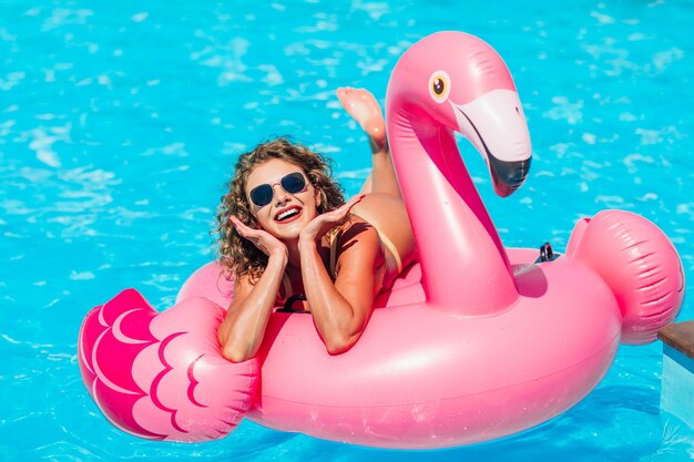 Blonde girlposing for instsgram stories, resting  in the  summer pool on an inflatable pink flamingo in a bathing suit.