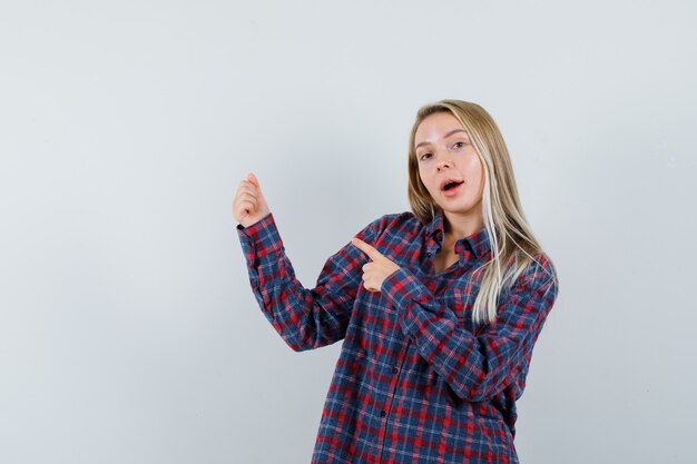 Blonde lady in casual shirt pointing at something pretended to be held and looking jolly , front view.