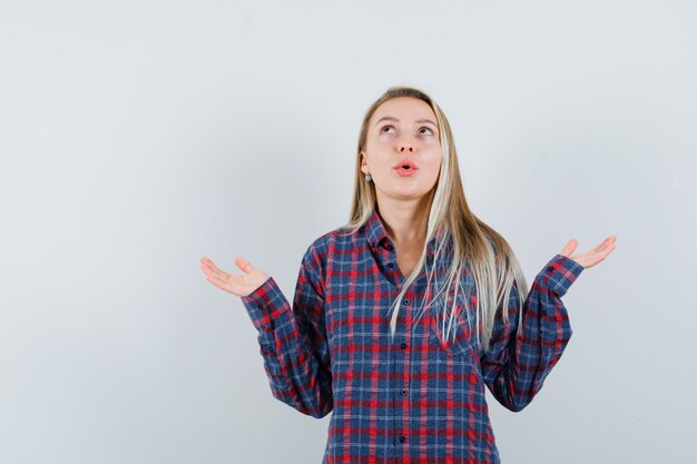 Blonde lady in casual shirt raising hands, looking up and looking amazed , front view.