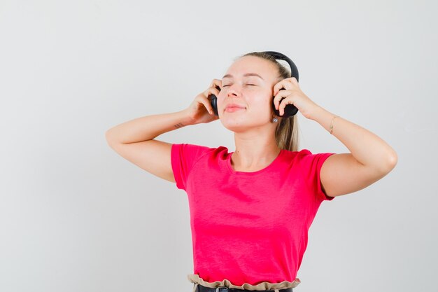 Blonde lady enjoying music with headphones in pink t-shirt and looking delighted , front view.