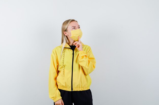 Blonde lady holding finger on chin in tracksuit, mask and looking pensive. front view.