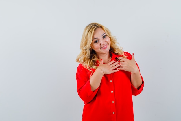 Blonde lady holding hand on her chest in red shirt and looking glad space for text