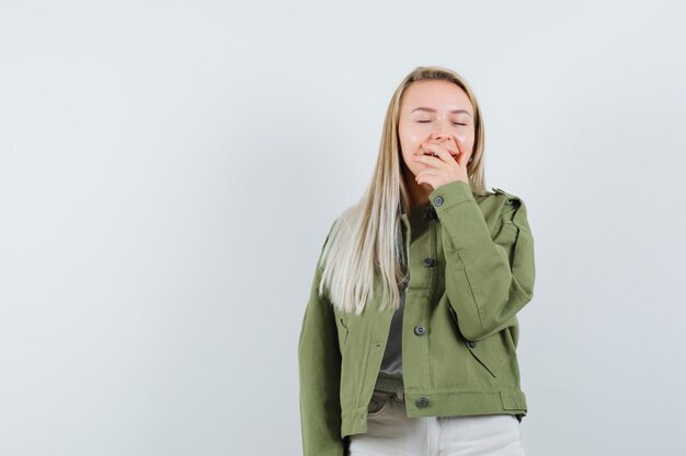 Blonde lady holding hand on mouth in jacket, pants and looking glad. front view.