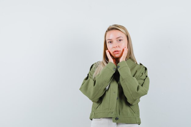 Blonde lady holding hands on cheeks in jacket, pants and looking elegant , front view.