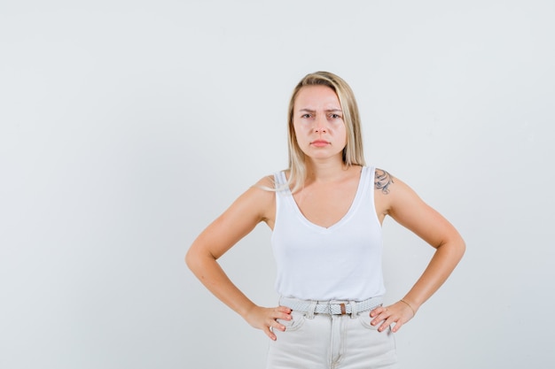 Blonde lady holding hands on waist in singlet, pants and looking spiteful. front view.