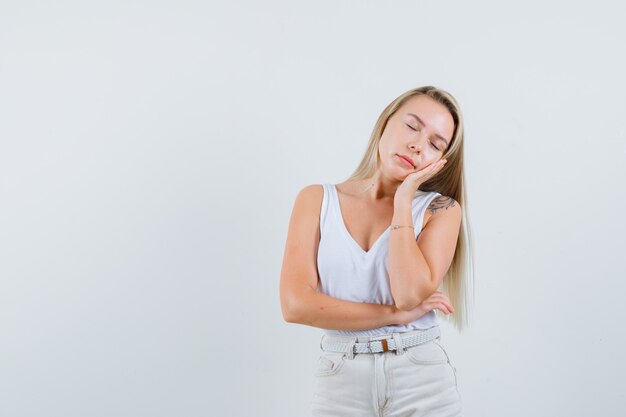 Blonde lady leaning cheek on raised palm in singlet, pants and looking sleepy