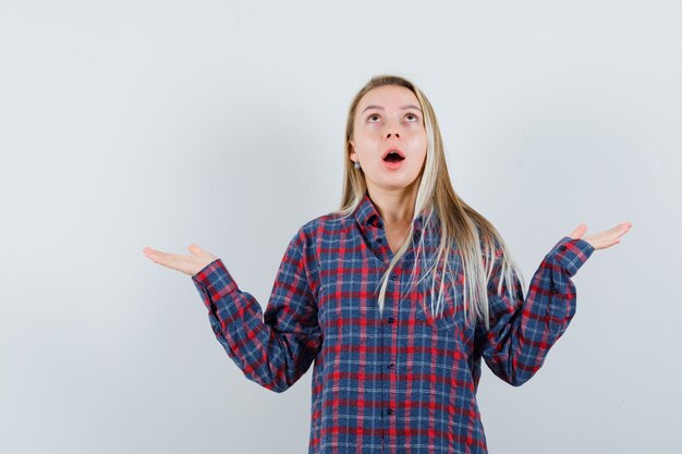 Blonde lady looking up while spreading palms in casual shirt and looking surprised , front view.