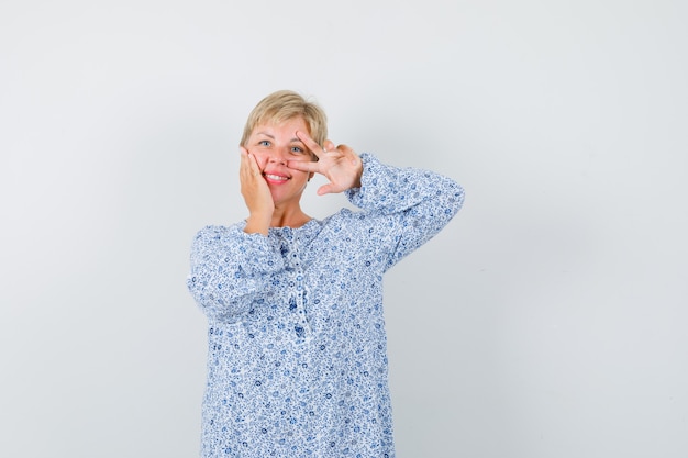 Blonde lady in patterned blouse showing v-sign over her eye and looking jolly , front view.