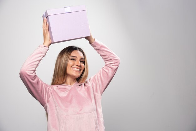 Blonde lady in pink sweatshirt holds a gift box over her head and shaking.