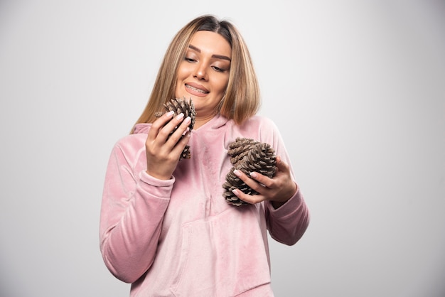 Free photo blonde lady in pink sweatshirt makes positive and happy face by holding oak tree cones.