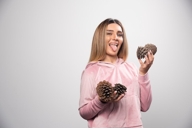 Free photo blonde lady in pink sweatshirt makes silly positive faces with oak tree cones in the hand.