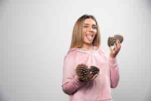 Free photo blonde lady in pink sweatshirt makes silly positive faces with oak tree cones in the hand.