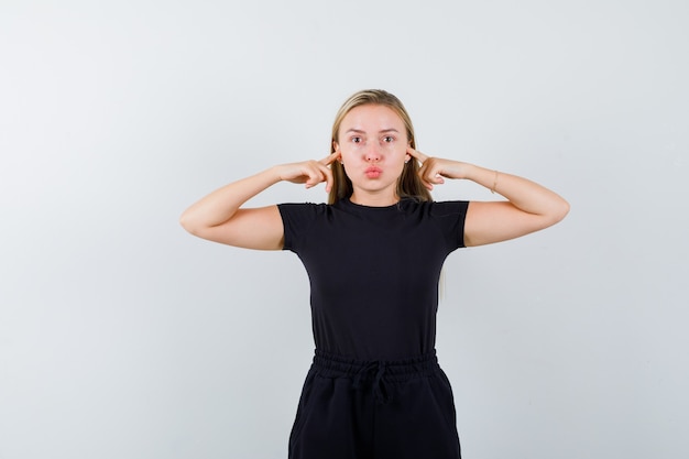 Free photo blonde lady plugging ears with fingers, pouting lips in black dress and looking careful. front view.