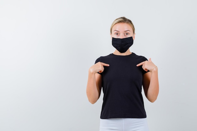 Blonde lady pointing at herself in black t-shirt, black mask and looking surprised isolated