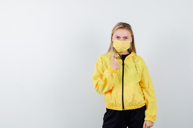 Blonde lady pointing at herself in tracksuit, mask and looking puzzled. front view.