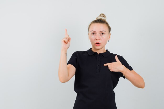 Blonde lady pointing at herself with finger up in black t-shirt and looking puzzled. front view.