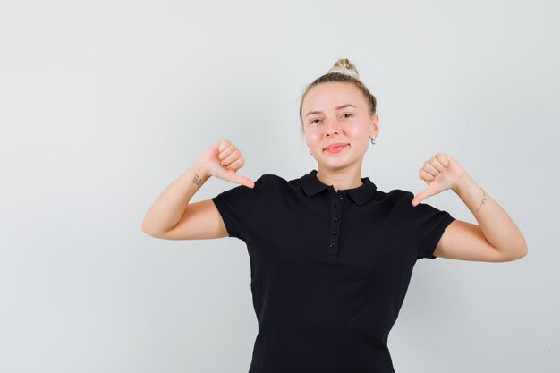 Blonde lady pointing at herself with thumbs in black t-shirt and looking confident , front view.