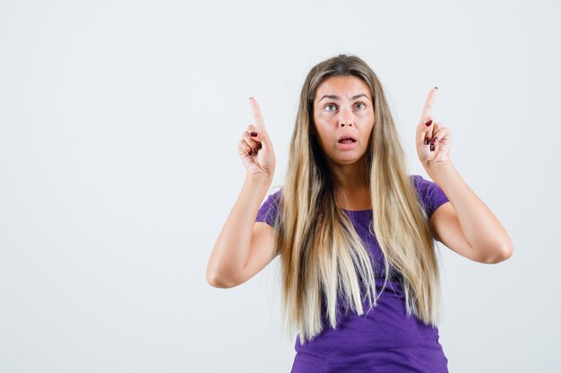 Blonde lady pointing up in violet t-shirt and looking puzzled. front view.