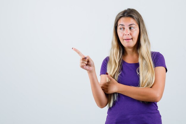 Blonde lady pointing at upper left corner in violet t-shirt front view.