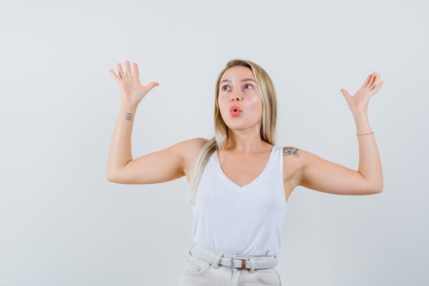 Blonde lady raising hands and looking up in singlet, pants and looking dreamy. front view.