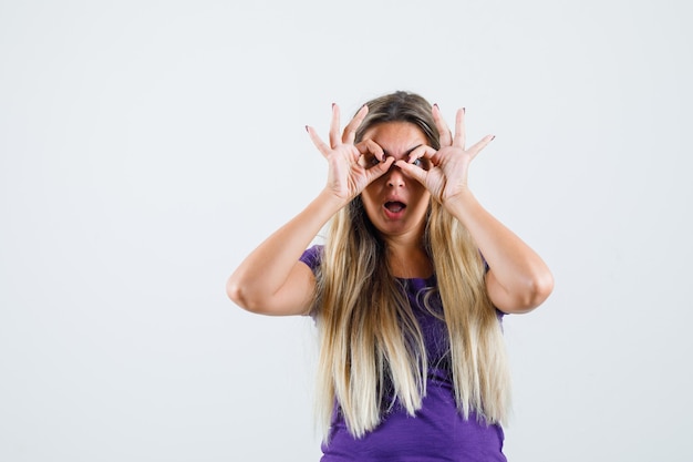 Blonde lady showing glasses gesture in violet t-shirt and looking amazed , front view.