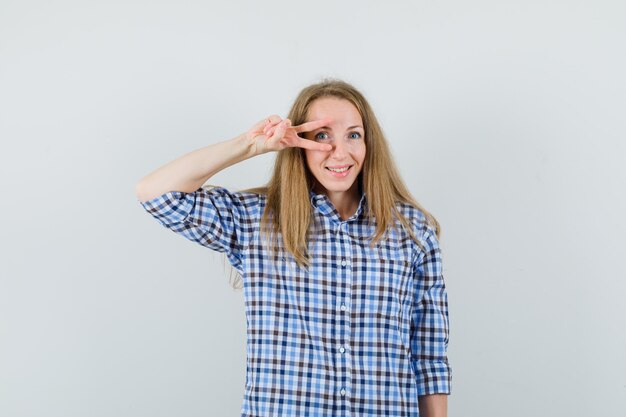 Free photo blonde lady showing v-sign on eye in shirt and looking cheerful.