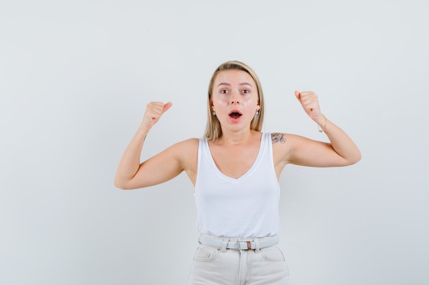 Blonde lady showing winner gesture in singlet, pants and looking lucky , front view.