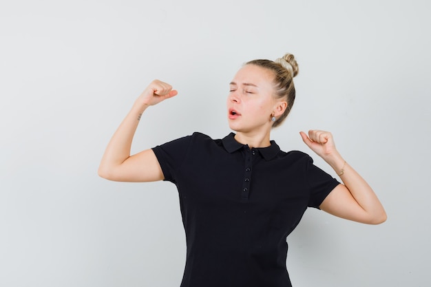 Blonde lady showing winner gesture with closed eyes in black t-shirt , front view.