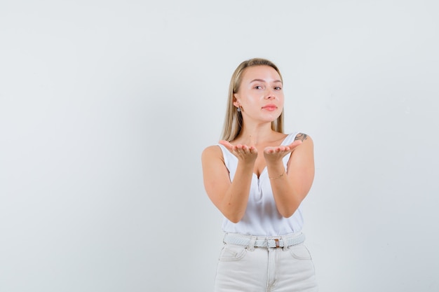 Blonde lady in singlet, pants making giving gesture and looking gentle , front view.