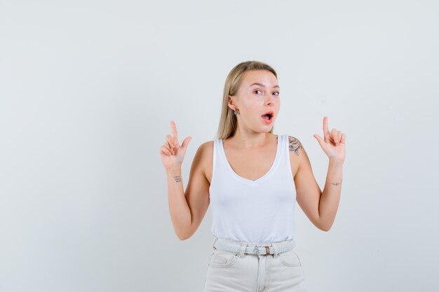 Blonde lady in singlet, pants pointing up and looking amazed , front view.