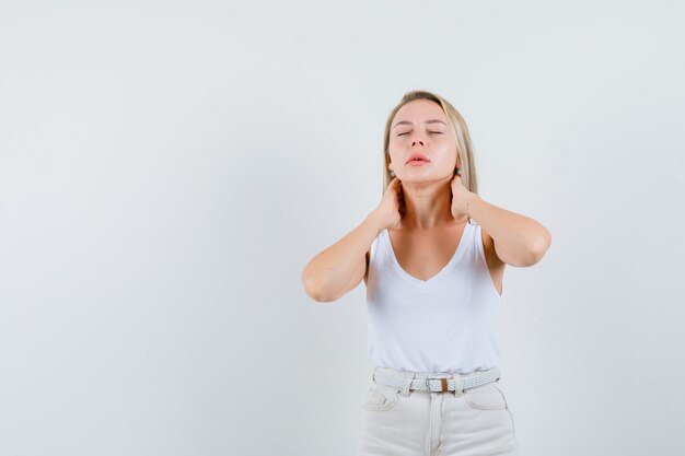 Blonde lady in singlet, pants suffering from neck pain and looking fatigued , front view.