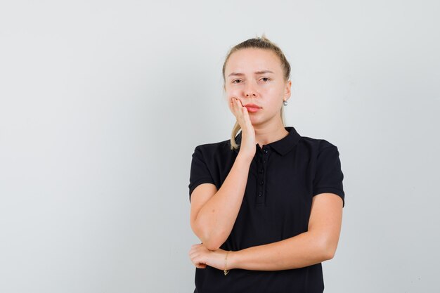 Blonde lady standing in thinking pose in black t-shirt and looking sad, front view.