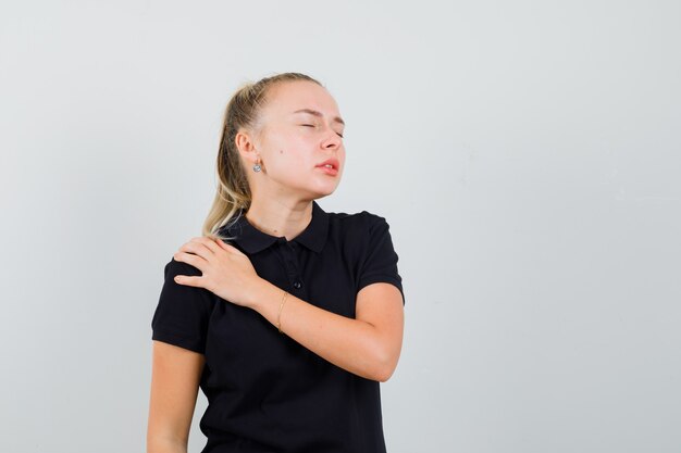 Blonde lady suffering from shoulder pain in black t-shirt and looking fatigued. front view.