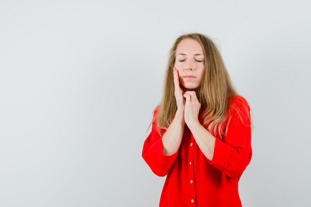 Blonde lady touching face skin on cheek in red shirt and looking calm.