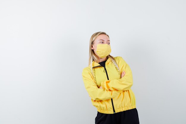 Blonde lady in tracksuit, mask holding arms folded and looking pensive , front view.