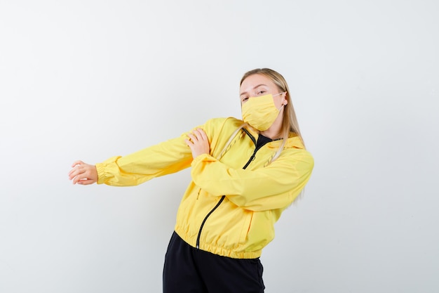 Free Photo blonde lady in tracksuit, mask posing while stretching arms and looking relaxed , front view.