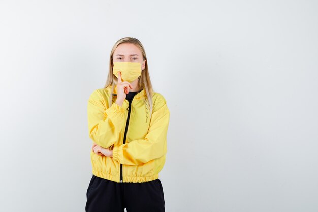 Blonde lady in tracksuit, mask standing in thinking pose and looking serious , front view.