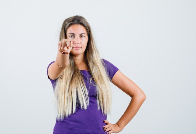 Blonde lady in violet t-shirt pointing at front and looking confident , front view.