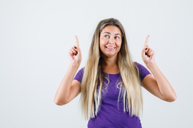 Blonde lady in violet t-shirt pointing up and looking cheerful , front view.