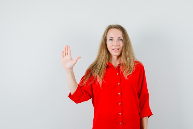 Blonde lady waving hand to say goodbye in red shirt and looking confident.