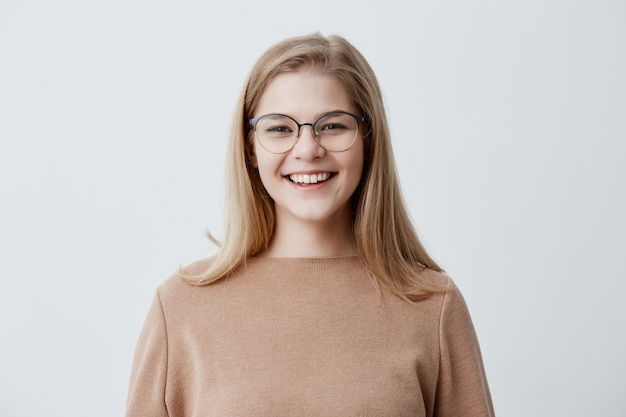 Blonde playful young European female wearing brown oversize sweater laughing at good joke, demonstrating perfect white teeth, her look and expression full of joy and happiness. People and lifestyle