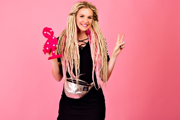 Blonde pretty woman with long blonde dreads holding plastic flamingo, wearing stylish sport chic outfit, surprised emotions, pop last style.