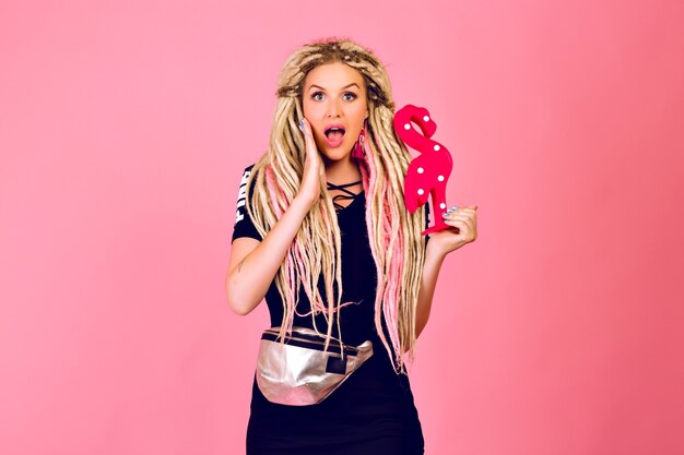 Blonde pretty woman with long blonde dreads holding plastic flamingo, wearing stylish sport chic outfit, surprised emotions, pop last style.