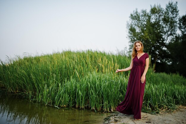 Blonde sensual barefoot woman in red marsala dress posing against lake with reeds