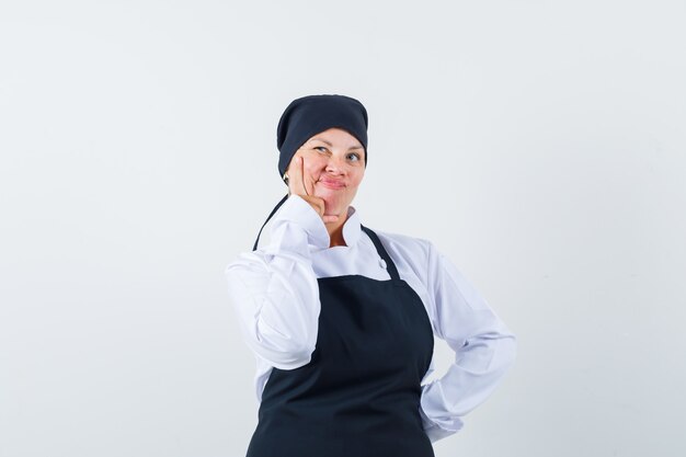 Blonde woman in black cook uniform holding index finger near mouth and looking pensive